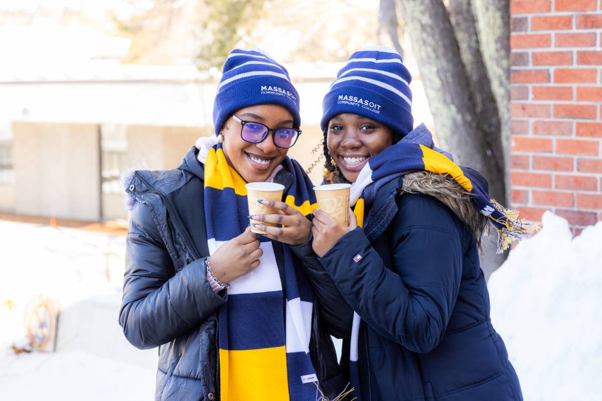Two students smiling at the camera while holding hot chocolate mugs and standing outside in the snow.