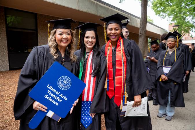 Three graduates posing outside while one holds a Liberal Arts Transfer Science sign.